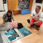 Városi Humánsegítő és Szociális Szolgálat | Elsősegélynyújtás kisgyermekkorban-szülőcsoportos megbeszélés a Bölcsődében Mentős tanít két embert stabil oldalfekvésre.