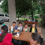 Városi Humánsegítő és Szociális Szolgálat | Napközis tábor a Családsegítőben Fiatalok étkeznek több kültéri padnál.