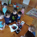 Kisgyerekek egy csoportja, akik szórakoztató húsvéti tojásfestéssel foglalkoznak egy fából készült asztal körül, figyelmük a színes tojásokra és a festékpoharakra összpontosul.