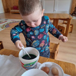 Egy kisgyerek óvatosan belemárt egy húsvéti tojást egy csésze zöld festékbe, és a tojásfestési tevékenység során figyelmesen a feladatra koncentrál.