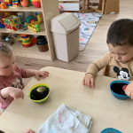 Két kisgyermek falatoznak egy kis asztalnál, tálakkal és színes játékokkal szétszórva a szobában, kiemelve a mindennapi gyermekkor egy pillanatát.