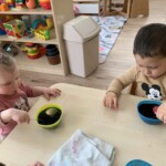 Városi Humánsegítő és Szociális Szolgálat | Húsvéti készülődés a bölcsődében. Két kisgyermek falatoznak egy kis asztalnál, tálakkal és színes játékokkal szétszórva a szobában, kiemelve a mindennapi gyermekkor egy pillanatát.