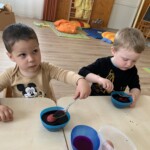 Városi Humánsegítő és Szociális Szolgálat | Húsvéti készülődés a bölcsődében. Két kisgyerek rágcsálnivalóra koncentrált egy gyerekméretű asztalnál egy játékokkal teli játszószobában.