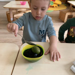 Egy fiatal gyerek, aki figyelmesen összpontosít, miközben megpróbál kikanalazni valamit egy zöld tálból egy gyerekméretű asztalnál.