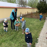Egy család élvezi a szabadtéri húsvéti tojásvadászatot, miközben egy felnőtt egy kosarat színes tojásokkal mutat meg a figyelmes gyerekeknek a kertben.
