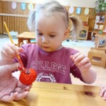 Városi Humánsegítő és Szociális Szolgálat | Húsvéti készülődés a bölcsődében. Egy kíváncsi copfos kisgyerek, aki feszülten fest egy piros tárgyat egy faasztalnál ülve, kreatív tevékenységbe merülve.