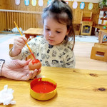 Fiatal gyerek koncentrált arckifejezéssel tojást fest ecsettel egy fából készült asztalnál, művészeti kellékekkel és színes tantermi környezettel a háttérben.