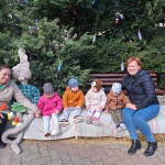 Két felnőtt és négy gyerek ül a szabadban egy ünnepileg feldíszített padon, nagy kitömött nyuszifigurákkal és szezonális dekorációkkal, és egy vidám pillanatot élveznek együtt.