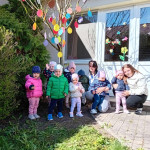 Gyermekcsoport a gondozóikkal, akik egy napsütéses napot élveznek a szabadban, egy színes húsvéti tojásdíszekkel díszített fa előtt állva.