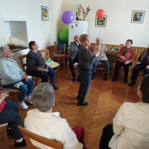 Városi Humánsegítő és Szociális Szolgálat | Galéria Körben ülő idősek középen egy ember táncol.