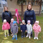 Városi Humánsegítő és Szociális Szolgálat | Húsvéti készülődés a bölcsődében. Vidám kisgyerekek csoportja, akik tanáraikkal vagy gondozóikkal a szabadban pózolnak fotózni egy napsütéses napon, mögöttük egy ünnepi szalaggal díszített szerkezet áll.