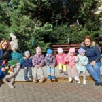 Városi Humánsegítő és Szociális Szolgálat | Húsvéti készülődés a bölcsődében. Két nő és egy csoport kisgyerek ülnek együtt kint egy padon, és élvezik a napsütéses napot zöldövezetben, körülöttük néhány játékos dekorációval.
