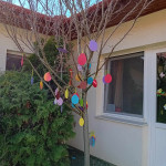 Színes húsvéti tojásdíszekkel díszített fa egy ház előtt, amely az ünnepi húsvéti ünnepséget jelzi.