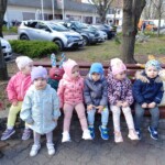 Városi Humánsegítő és Szociális Szolgálat | Húsvéti készülődés a bölcsődében. Egy csoport kisgyerek ül egymás mellett a szabadban egy padon, színes tavaszi kabátot és sapkát visel, parkolóval és fákkal a háttérben.