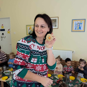 Egy nő tart a kezében egy kézzel készített díszt, háttérben gyerekek egy nagy asztalnál