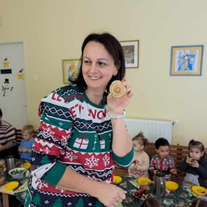 Egy nő tart a kez&eacute;ben egy k&eacute;zzel k&eacute;sz&iacute;tett d&iacute;szt, h&aacute;tt&eacute;rben gyerekek egy nagy asztaln&aacute;l